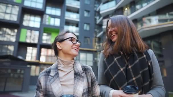 Twee gelukkige vrouwen wandelen met afhaalkoffie en praten met interesse onder elkaar in het zakendistrict. Langzame beweging — Stockvideo