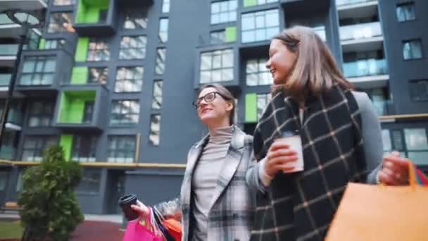 Dos mujeres felices caminan con bolsas de compras y café para llevar después de una compra exitosa y hablan con interés entre ellas. — Vídeo de stock