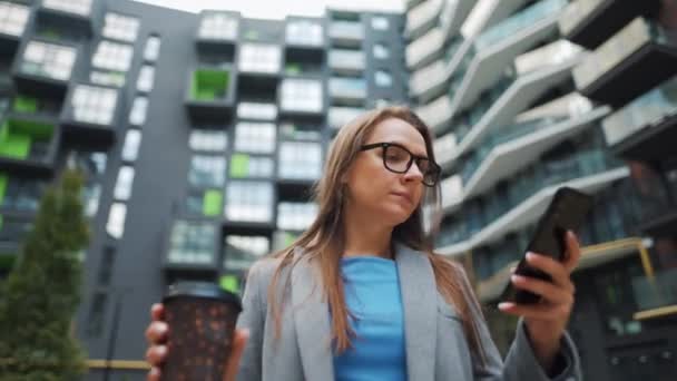 Wanita berpakaian formal berjalan menyusuri jalan di daerah bisnis dengan kopi di tangan dan menggunakan smartphone — Stok Video