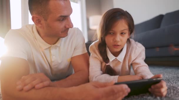 Отец и дочь наслаждаются временем вместе и с помощью планшета для семейного развлечения, лежа на полу в гостиной на дому. — стоковое видео