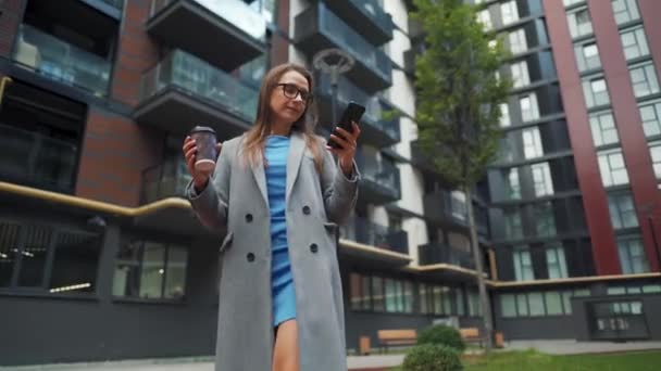 Formell gekleidete Frau läuft mit Kaffee in der Hand und Smartphone durch ein Geschäftsviertel — Stockvideo