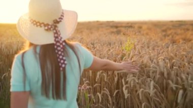 Şapkalı ve mavi elbiseli bir kadın buğday tarlası boyunca yürüyor ve gün batımında eli ile olgunlaşmış buğday başaklarına dokunuyor. Yavaş çekim