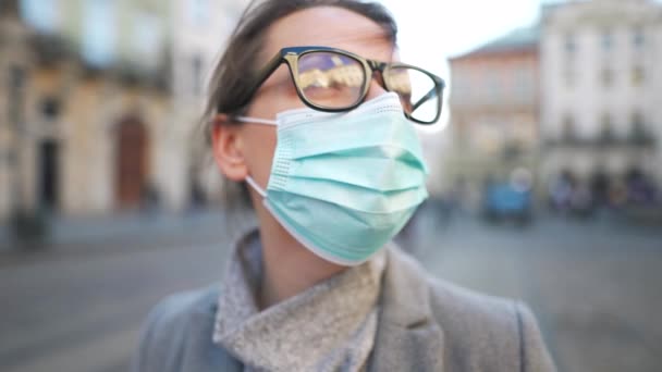 Pandemische bescherming van het Covid-19 coronavirus. Portret van een vrouw in een jas, bril en een beschermend medisch masker. Ze staat midden op het plein. Langzame beweging — Stockvideo