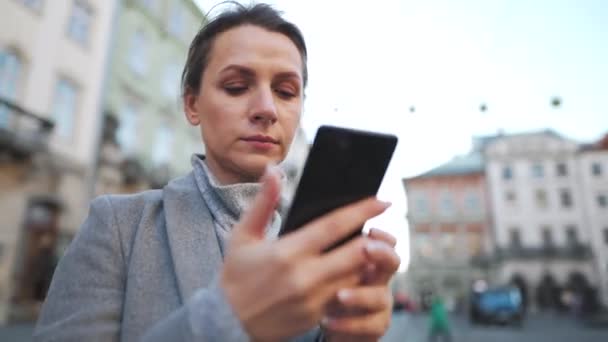 Žena v kabátě stojí uprostřed starého náměstí, používá smartphone a fotí — Stock video