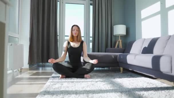 Siyah tulumlu genç beyaz kadın lotus pozisyonunda oturuyor ve meditasyon yapıyor, yoga yapıyor.. — Stok video