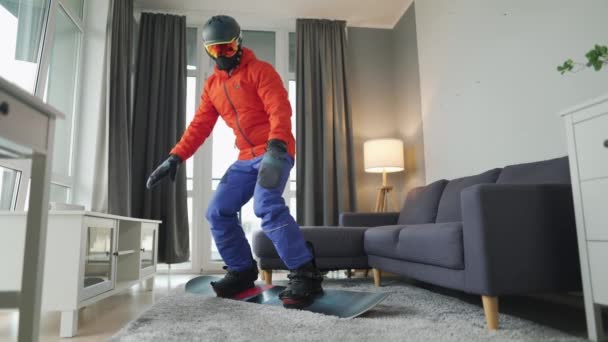 Vídeo divertido. Hombre vestido de snowboarder monta una tabla de snowboard en una alfombra en una habitación acogedora. Esperando un invierno nevado — Vídeos de Stock