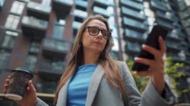 Resmi olarak giyinmiş bir kadın elinde termo bardakla bir iş bölgesinde duruyor ve akıllı telefon kullanıyor. Kamera onun etrafında dönüyor.