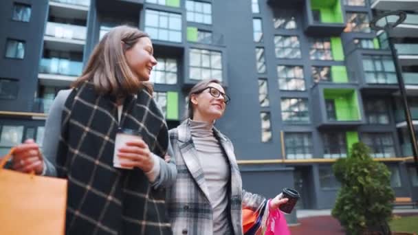Twee gelukkige vrouwen lopen met boodschappentassen en afhaalkoffie na een succesvol winkelen en praten met interesse onder elkaar. Langzame beweging — Stockvideo