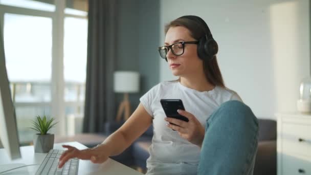 Mujer vestida casualmente con auriculares que trabajan con una computadora y un teléfono inteligente en casa en un ambiente acogedor. Trabajo remoto fuera de la oficina. — Vídeo de stock