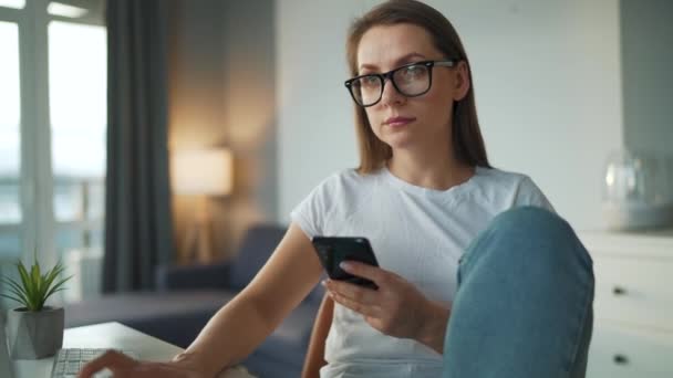 Casually dressed woman working with a computer and smartphone at home in a cozy environment. Remote work outside the office. — Stock Video