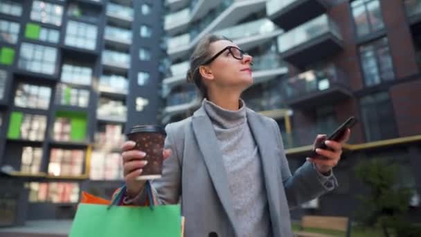 Vrouw loopt op een zakenwijk, houdt afhaalkoffie, boodschappentassen en maakt gebruik van smartphone. Stedelijke levensstijl en digitale technologie — Stockvideo