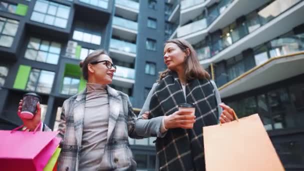 Dos mujeres felices caminan con bolsas de compras y café para llevar después de una compra exitosa y hablan con interés entre ellas — Vídeos de Stock