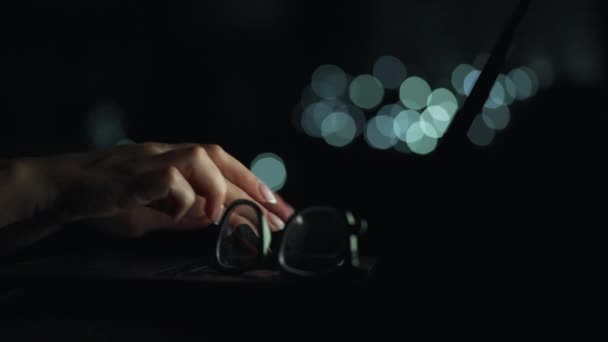 Ženský ruce píšou v noci na notebook. Brýle jsou na popředí. Rozmazaná světla na pozadí — Stock video