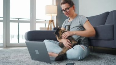 Sıradan giyimli bir kadın dizüstü bilgisayarla halıya oturur, dizlerinin üzerine çöker ve tüylü bir kediyi okşar ve rahat bir odada çalışır. Ofis dışında uzaktan çalışma.