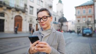 Gözlüklü bir kadın eski bir caddede yürürken palto giyiyor ve akıllı telefon kullanıyor. Arka planda sarı tramvay geçiyor..