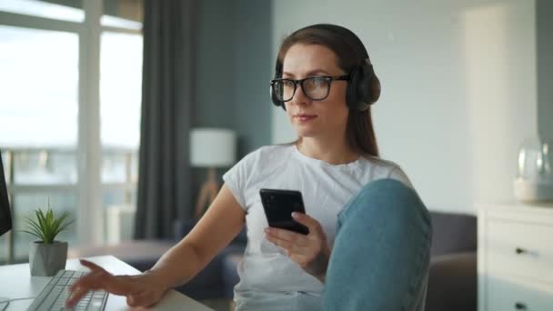 Mujer vestida casualmente con auriculares que trabajan con una computadora y un teléfono inteligente en casa en un ambiente acogedor. Trabajo remoto fuera de la oficina. — Vídeo de stock