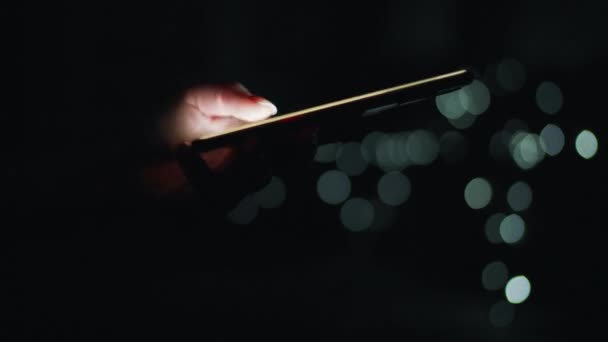 Weibliche Hände tippen nachts auf einem Smartphone. Verschwommenes Licht auf dem Hintergrund — Stockvideo