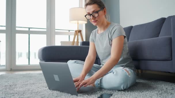 Lässig gekleidete Frau sitzt auf Teppich mit Laptop und Smartphone und arbeitet in gemütlichem Raum. Fernarbeit außerhalb des Büros. — Stockvideo