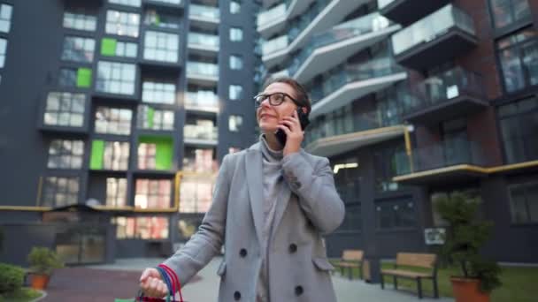 Kvinna promenader på ett affärsdistrikt med shoppingväskor och prata på smartphone. Stadsliv och digital teknik — Stockvideo