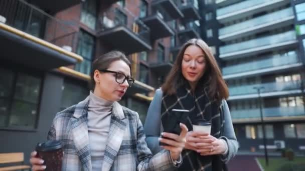 Twee gelukkige vrouwen wandelen met afhaalkoffie en praten met interesse onder elkaar in de zakenwijk. — Stockvideo