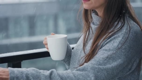 Mulher fica na varanda durante a queda de neve com xícara de café quente ou chá. Ela olha para os flocos de neve e respira no ar fresco gelado. — Vídeo de Stock