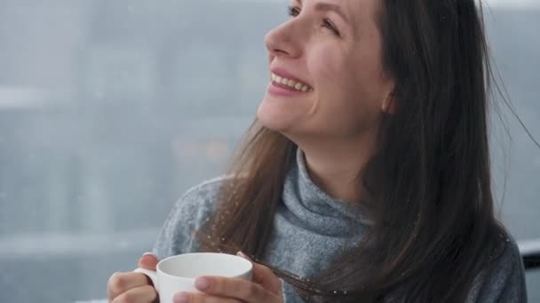 Mulher caucasiana fica na varanda durante a queda de neve com xícara de café quente ou chá. Ela olha para os flocos de neve e respira no ar fresco gelado — Vídeo de Stock