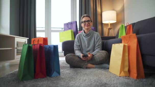 Porträt einer glücklichen Frau, die auf einem Teppich in einem gemütlichen Raum zwischen Einkaufstüten sitzt — Stockvideo