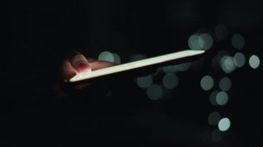 Kadın eli, gece akıllı telefonunda daktilo çalıyor. Arkaplanda bulanık ışıklar