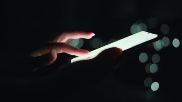 A női kezek éjszaka egy okostelefonon gépelnek. Homályos fények a háttérben — Stock videók