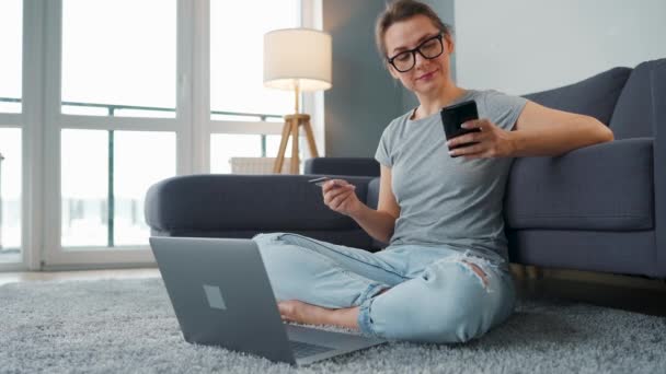 Mujer con gafas se sienta en el suelo y hace una compra en línea con una tarjeta de crédito y un teléfono inteligente. Compras en línea, tecnología de estilo de vida — Vídeos de Stock