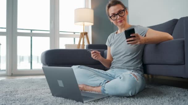 Frau mit Brille sitzt auf dem Boden und kauft online mit Kreditkarte und Smartphone ein. Online-Shopping, Lifestyletechnologie — Stockvideo