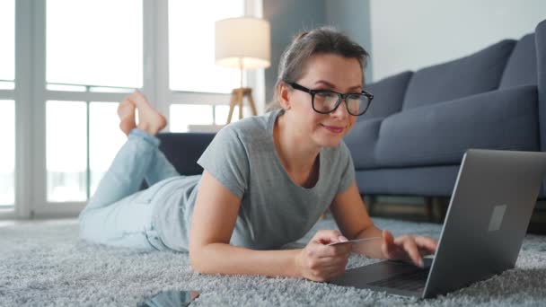 Kobieta w okularach leży na podłodze i dokonuje zakupu online za pomocą karty kredytowej i smartfona. Zakupy online, technologia stylu życia — Wideo stockowe