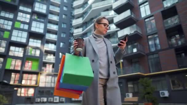 Kvinna promenader på ett affärsdistrikt, rymmer hämtmat kaffe, shoppingväskor och använda smartphone. Stadsliv och digital teknik — Stockvideo