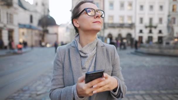 Een vrouw in een jas die door de straat loopt, een smartphone gebruikt en een foto maakt. Langzame beweging — Stockvideo