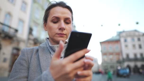 Žena v kabátě, která stojí uprostřed starého náměstí, používá smartphone a fotí se. Zpomalený pohyb — Stock video