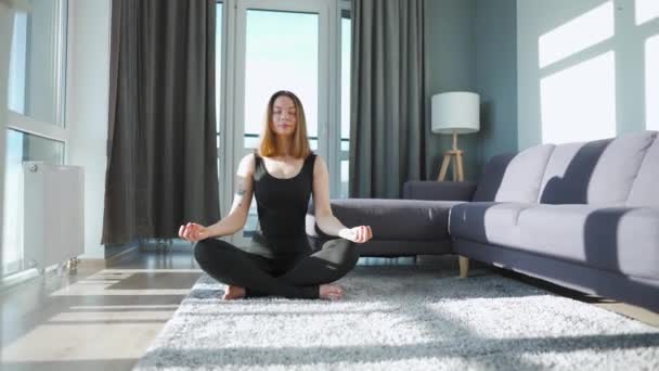 Ung kaukasisk kvinna i svart overall sitter i lotusställning och mediterar, yoga hemma. Långsamma rörelser — Stockvideo