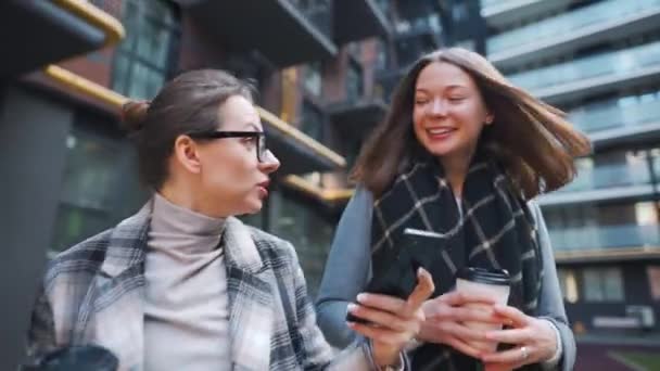 Zwei glückliche Frauen spazieren mit Kaffee zum Mitnehmen und unterhalten sich interessiert untereinander im Geschäftsviertel. — Stockvideo