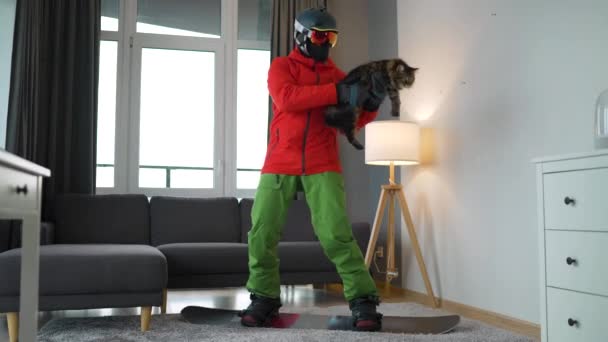 Весёлое видео. Человек, одетый сноубордистом, катается на сноуборде по ковру в уютной комнате. Он держит пушистую кошку на руках. В ожидании снежной зимы — стоковое видео