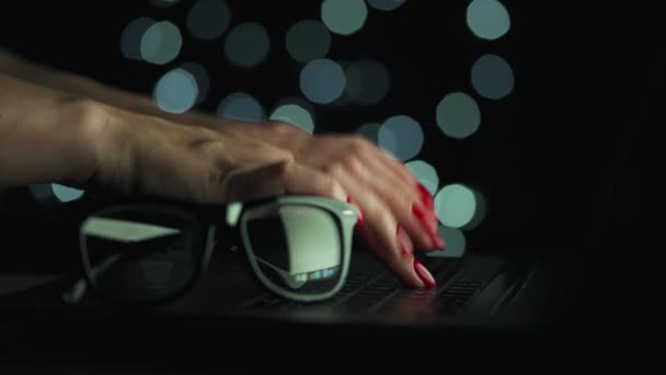 Las manos femeninas están escribiendo en un portátil por la noche. Las gafas están en primer plano. Luces borrosas en el fondo — Vídeo de stock