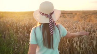 Şapkalı ve mavi elbiseli bir kadın buğday tarlası boyunca yürüyor ve gün batımında eli ile olgunlaşmış buğday başaklarına dokunuyor. Yavaş çekim