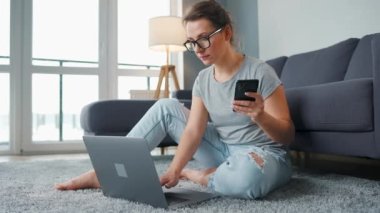 Halıda dizüstü bilgisayarı ve akıllı telefonu olan rahat bir odada çalışan sıradan giyinmiş bir kadın. Ofis dışında uzaktan çalışma.