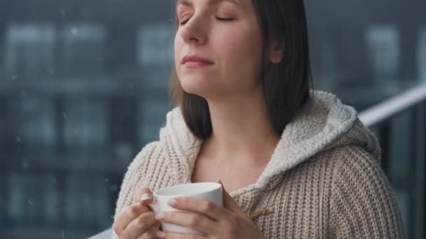 Kobieta pozostaje na balkonie podczas opadów śniegu z filiżanką gorącej kawy lub herbaty. Patrzy na płatki śniegu i oddycha mroźnym świeżym powietrzem.. — Wideo stockowe
