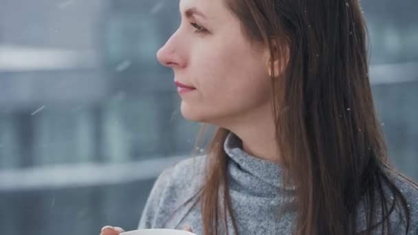 女性は熱いコーヒーか紅茶のコップとの降雪の間バルコニーにとどまる。彼女は雪の結晶を見て、新鮮な空気の中で息をする. — ストック動画