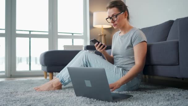 Mujer vestida casualmente sentada en la alfombra con portátil y teléfono inteligente y trabajando en una habitación acogedora. Trabajo remoto fuera de la oficina. — Vídeos de Stock