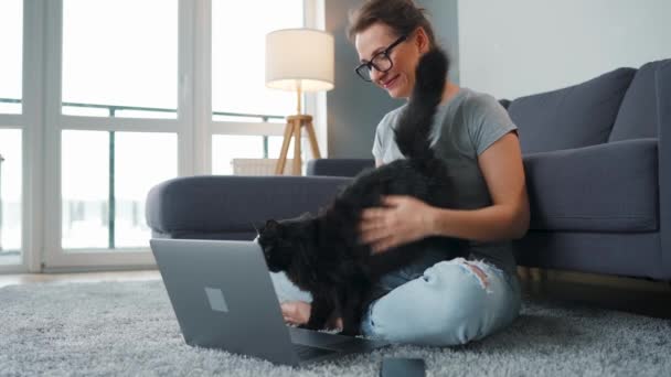 Mulher casualmente vestida com óculos senta-se em um tapete com um laptop andstrokes um gato preto fofo, enquanto trabalha em um quarto acolhedor. — Vídeo de Stock