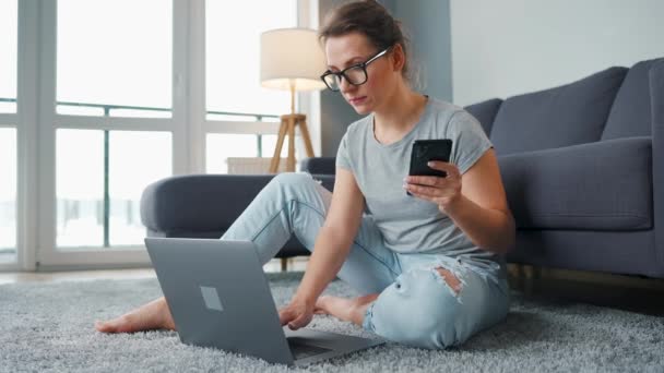 Mujer vestida casualmente sentada en la alfombra con portátil y teléfono inteligente y trabajando en una habitación acogedora. Trabajo remoto fuera de la oficina. — Vídeos de Stock