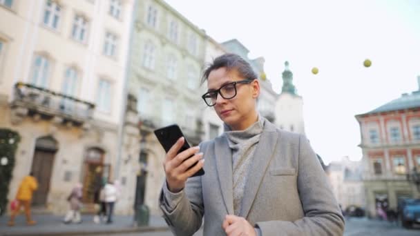 Mujer con gafas que lleva un abrigo caminando por una calle vieja y usando un smartphone. Movimiento lento — Vídeos de Stock