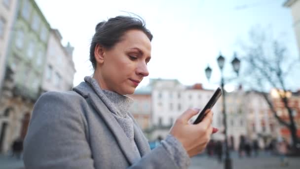 Žena v kabátě, která stojí uprostřed starého náměstí, používá smartphone a fotí se. Zpomalený pohyb — Stock video