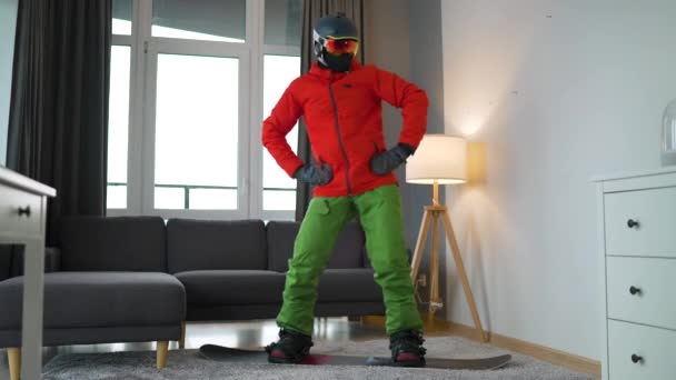 Vidéo amusante. Homme habillé comme un snowboarder dansant joyeusement et s'amusant tout en se tenant sur un snowboard sur un tapis dans une salle confortable. En attendant un hiver enneigé — Video