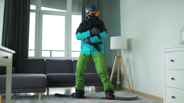 Vídeo divertido. Hombre vestido de snowboarder monta una tabla de snowboard en una alfombra en una habitación acogedora. Tiene un gato esponjoso en sus brazos. Esperando un invierno nevado. Movimiento lento — Vídeo de stock